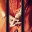 A person in an orange prison outfit with handcuffs standing behind bars. A person in an orange prison outfit with handcuffs standing behind bars.