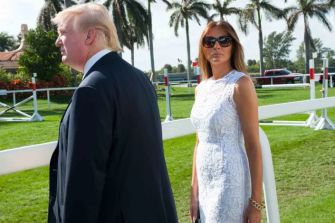 Two individuals outdoors, with palm trees in the background; one wearing sunglasses and a white dress, while the other is dressed in a suit.