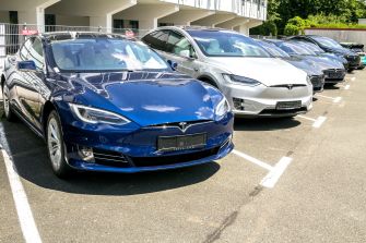 A row of Tesla electric vehicles parked in a lot, including a blue Tesla Model S and a silver Tesla Model X.