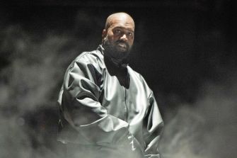 Kanye West performing on stage, dressed in a stylish black outfit with a serious expression amid fog and lighting.