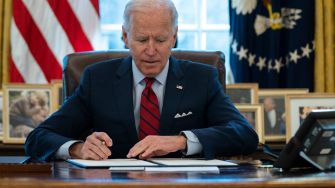 President Biden signing documents at a desk in the Oval Office, with American flags and family photos in the background.