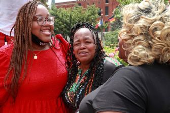 Women embracing and smiling together in a park, with flowers and colorful flags in the background.