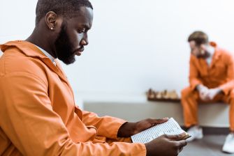 A man in an orange prison jumpsuit reading a publication while another inmate sits in the background.
