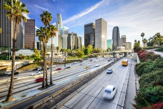 A bustling highway scene in Los Angeles with skyscrapers in the background and vehicles driving under a clear blue sky.
