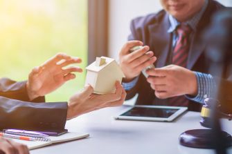 Two professionals discussing estate planning, with one holding a miniature house and the other handling cash. A gavel and notepad are visible on the table.