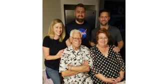 A family gathering with five individuals, including an elderly man seated in front, smiling with his arms crossed, surrounded by younger family members.