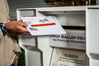 Person placing an official ballot envelope into a drop box labeled "Drop Ballot Here" with a warning sign about tampering.