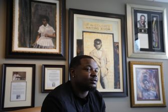 A man seated in an office surrounded by framed artwork and photographs, reflecting on his experiences related to a wrongful arrest.