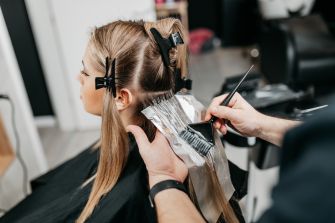 A woman getting her hair dyed in a salon, with foils in her hair and a stylist applying color.