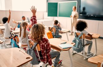 A classroom scene with students raising their hands while a teacher addresses the class.