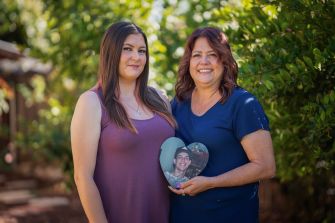 A mother and daughter holding a heart-shaped photo frame, featuring a picture of a young man, outdoors in a garden setting.