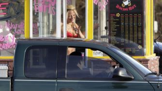 A barista in a bikini at a drive-thru coffee stand interacts with a customer in a vehicle.