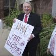 A man holding a sign that reads "FREE the Electors" outside a courthouse, surrounded by supporters. A man holding a sign that reads "FREE the Electors" outside a courthouse, surrounded by supporters.