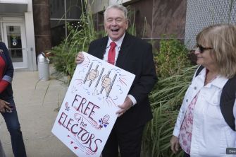 A man holding a sign that reads "FREE the Electors" outside a courthouse, surrounded by supporters.