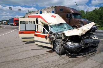 Damaged ambulance involved in a serious accident on a highway, with trucks in the background.