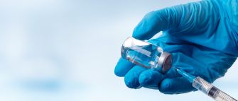 A hand in a blue glove holding a vaccine vial with a syringe poised to draw the vaccine.