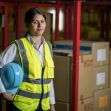 A woman in a safety vest and hard hat stands in a warehouse surrounded by cardboard boxes. A woman in a safety vest and hard hat stands in a warehouse surrounded by cardboard boxes.