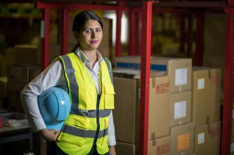 A woman in a safety vest and hard hat stands in a warehouse surrounded by cardboard boxes.