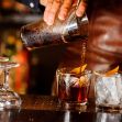 A bartender pouring a mixed drink into two glasses with garnishes at a bar. A bartender pouring a mixed drink into two glasses with garnishes at a bar.
