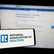 A person holding a smartphone displaying the National Association of Realtors (NAR) logo, with a computer screen showing real estate market trends in the background. A person holding a smartphone displaying the National Association of Realtors (NAR) logo, with a computer screen showing real estate market trends in the background.