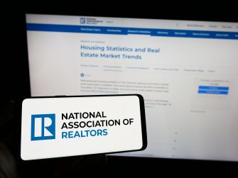 A person holding a smartphone displaying the National Association of Realtors (NAR) logo, with a computer screen showing real estate market trends in the background.