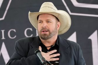 Garth Brooks speaking at a public event, wearing a cowboy hat and suit, with a serious expression.