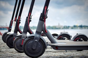 Electric scooters lined up in a row, set against a cloudy sky and waterfront background.