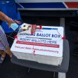 A person placing ballots into an official ballot box labeled "INSERT BALLOTS HERE" in a parking lot setting. A person placing ballots into an official ballot box labeled "INSERT BALLOTS HERE" in a parking lot setting.