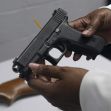 A person holding a Glock handgun, with a wooden firearm visible on a table in the background. A person holding a Glock handgun, with a wooden firearm visible on a table in the background.