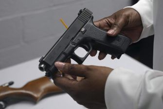 A person holding a Glock handgun, with a wooden firearm visible on a table in the background.