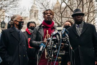 A group of individuals, including a man with a trumpet, speaking at a press conference in front of a building, addressing issues related to a viral incident involving racial profiling.