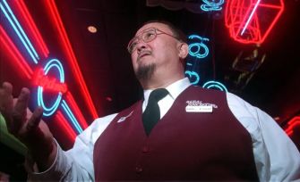 A man in a vest and glasses stands in a brightly lit venue with neon lights, gesturing as he speaks.