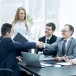 A group of professionals in business attire engaged in a meeting, with one man shaking hands with another across a conference table. A group of professionals in business attire engaged in a meeting, with one man shaking hands with another across a conference table.