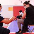 A healthcare worker assists a teenager receiving a vaccine in a gymnasium setting, both wearing masks. A healthcare worker assists a teenager receiving a vaccine in a gymnasium setting, both wearing masks.
