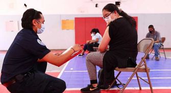 A healthcare worker assists a teenager receiving a vaccine in a gymnasium setting, both wearing masks.
