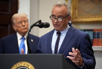 Robert F. Kennedy Jr. speaks at a podium in front of a Presidential seal, with a figure in the background.