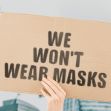 A person holding a cardboard sign that reads "WE WON'T WEAR MASKS" in front of a cityscape background. A person holding a cardboard sign that reads "WE WON'T WEAR MASKS" in front of a cityscape background.