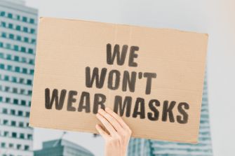 A person holding a cardboard sign that reads "WE WON'T WEAR MASKS" in front of a cityscape background.