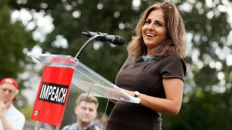 A woman speaking at a podium with a sign that reads "IMPEACH" in front of a blurred crowd.