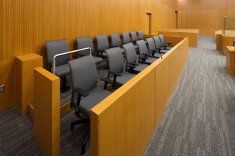 Image of an empty jury box inside a courtroom, featuring rows of gray chairs and wooden walls.