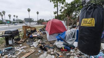 A homeless encampment cluttered with belongings and debris near a highway in California.