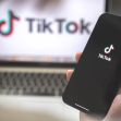A person holding a smartphone displaying the TikTok logo, with a laptop featuring the same logo in the background. A person holding a smartphone displaying the TikTok logo, with a laptop featuring the same logo in the background.