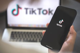 A person holding a smartphone displaying the TikTok logo, with a laptop featuring the same logo in the background.