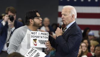 A scene depicting President Biden engaging with a protester holding a sign that reads "NOT 1 MORE DEPORTATION" during an event, amidst a gathering of supporters in the background.