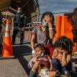 A mother and her two children sitting on the ground, visibly distressed, with police officers in the background during a border enforcement situation. A mother and her two children sitting on the ground, visibly distressed, with police officers in the background during a border enforcement situation.
