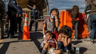 A mother and her two children sitting on the ground, visibly distressed, with police officers in the background during a border enforcement situation.