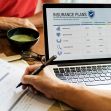 A person reviewing insurance plans on a laptop while filling out paperwork at a table with a bowl and calculator. A person reviewing insurance plans on a laptop while filling out paperwork at a table with a bowl and calculator.