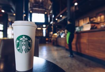 A Starbucks cup in the foreground with a blurred barista in the background at a Starbucks store.
