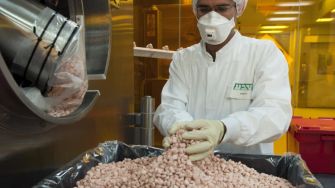 An employee in a lab coat and mask handles a large quantity of pharmaceutical pills in a manufacturing facility.