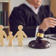 Wooden figures representing a family stand beside a judge's gavel on a table, emphasizing a child custody case in a courtroom setting. Wooden figures representing a family stand beside a judge's gavel on a table, emphasizing a child custody case in a courtroom setting.